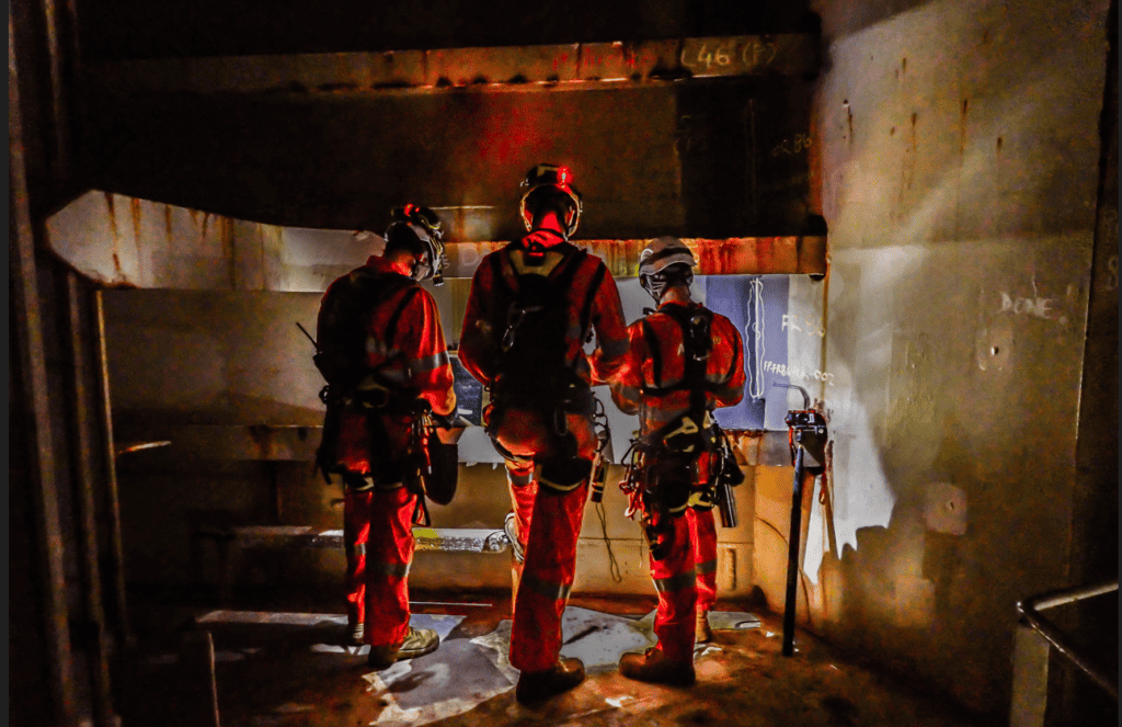 Three men wearing overalls, helmets and climbing gear, standing inside a cargo tank.